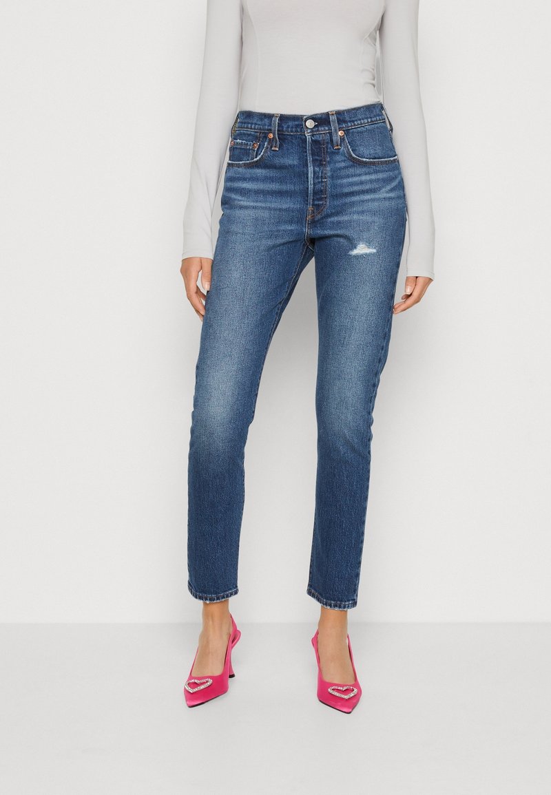 Jean en denim bleu à taille haute, jambes droites, légère usure au niveau de la cuisse, avec un design classique à cinq poches.
