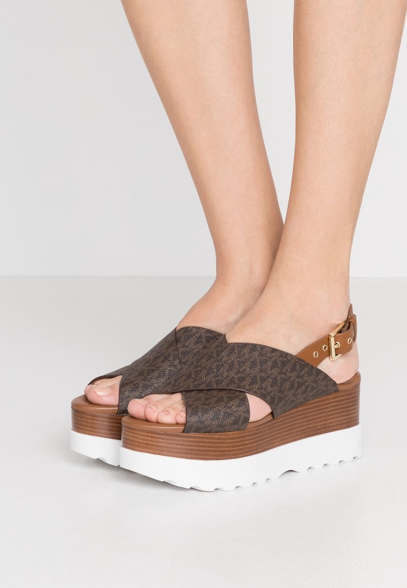 Sandales à talon ouvert avec un dessus texturé marron, des brides croisées et une boucle ajustable à la cheville. Dispose d'une plateforme marron imitant le bois et d'une semelle blanche.