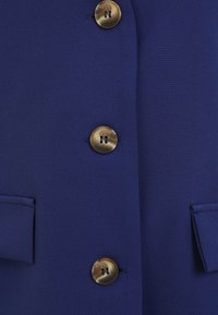 Blaue Jacke aus strukturiertem Stoff, mit zwei großen runden braunen Knöpfen und zwei Klapptaschen. Nahaufnahme der Knopfdetails und des Stoffes.