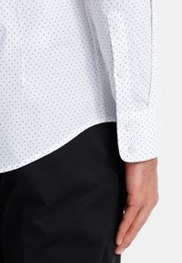 Camisa blanca de vestir con un pequeño patrón de puntos verdes, de mangas largas, con puños de botones, de material de algodón, diseño ajustado, dobladillo inferior curvado, combinada con pantalones oscuros.