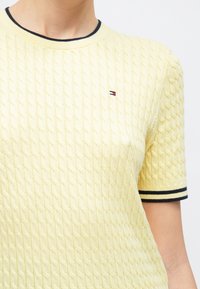 Κίτρινο πουλόβερ με πλέξη καλωδίου και κοντά μανίκια, με μπλε σκούρο ριμπ λαιμόκοψη και μανσέτες, και μικρό λογότυπο στην πάνω αριστερή γωνία.