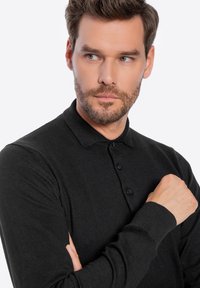 Schwarzes Langarm-Polohemd mit geripptem Kragen, drei Knöpfen und strukturiertem Stoff. Das Modell hat die Arme verschränkt, um die Passform des Shirts zu zeigen.