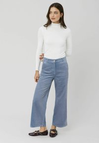 Pantalons larges en velours côtelé bleu clair avec des cannelures texturées, associés à un haut ajusté à manches longues blanc et des chaussures plates sombres.
