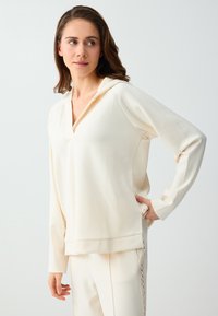 Femme aux cheveux bruns portant un haut à capuche crème à manches longues et un pantalon assorti avec un détail en dentelle sur le côté, regardant vers sa gauche.