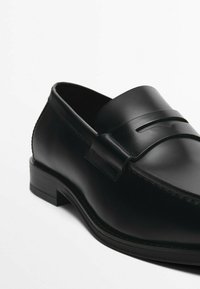 Schwarze Leder-Loafers mit glatter Oberfläche, ausgestattet mit einem dekorativen Riemen und einem Stichdetail sowie einem niedrigen rechteckigen Absatz.