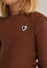 Top marrón de manga larga con relieve que presenta un pequeño logo bordado de un corazón en azul y blanco en el lado izquierdo del pecho.