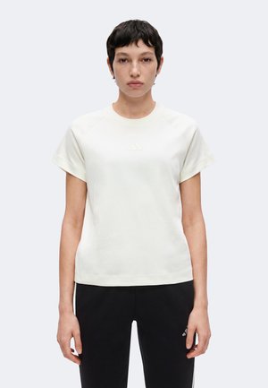 Personne avec des cheveux courts et foncés portant un t-shirt blanc uni à manches courtes et un pantalon noir, debout devant un fond clair uni.