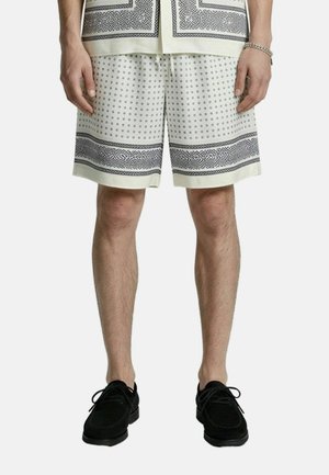 Man draagt witte short met zwarte stippen en randpatronen, zwarte veterschoenen en een bijpassend patroonhemd zichtbaar aan de zoom.