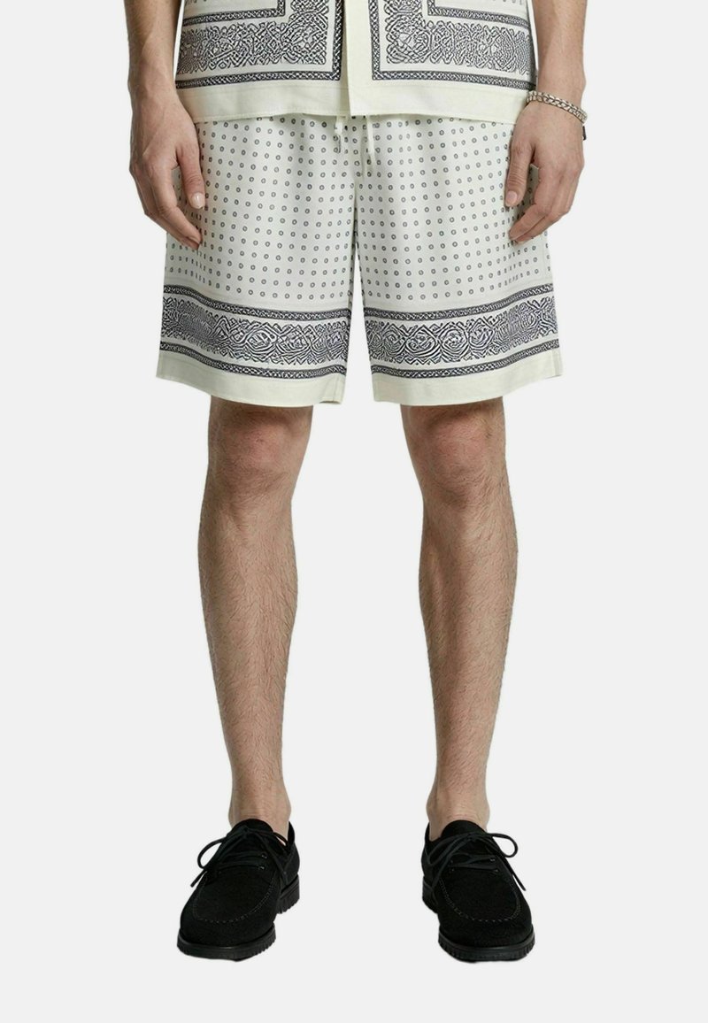 Hombre que lleva pantalones cortos blancos con patrones de puntos negros y bordes, zapatos negros con cordones y una camisa con un patrón a juego visible en el dobladillo.