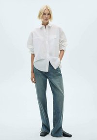 Camicia bianca a maniche lunghe realizzata in tessuto leggero, con tasca e vestibilità rilassata, abbinata a jeans a gamba larga blu chiaro e scarpe nere.