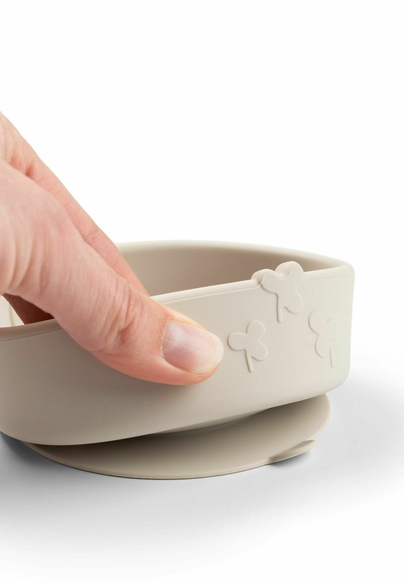 Main tenant un bol en silicone beige avec base ventouse et motifs en relief en forme de papillon sur le côté, sur fond blanc.