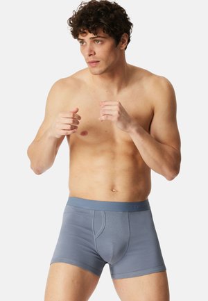 Skjorteløs mand i grå boxershorts står med hænderne hævet og ser opmærksomt til siden mod en ensfarvet baggrund.