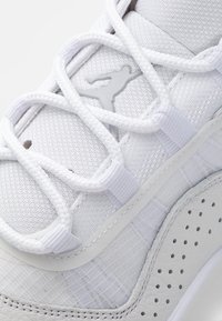Gros plan d'une basket blanche avec tissu texturé, lacets style corde, cuir perforé et un logo en relief en silhouette sur la languette.