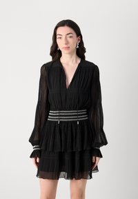 Black pleated dress with long sleeves, v-neck, and tiered ruffle hem. Features a silver-accented waistline band with a zigzag pattern.