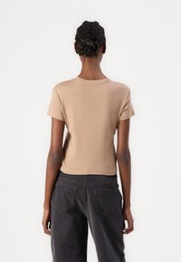 Tan short-sleeve top with a round neckline; fitted cropped style. Smooth texture with a subtle finish, paired with dark gray trousers featuring back pockets.