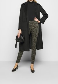 Abrigo negro largo sobre un cuello de tortuga negro, combinado con pantalones de estampado leopardo verde oliva y tacones puntiagudos negros. Clutch negro con borlas.