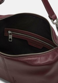 Wnętrze bordowej skórzanej torby z czarną podszewką. Zawiera kieszeń na zamek oraz metkę z logo marki. Gładka faktura z metalicznym zamkiem.