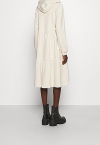 Robe à capuche crème avec un design à jupe à volants, manches longues et taille froncée ; associée à des bottines noires à la cheville. Texture de tissu lisse.