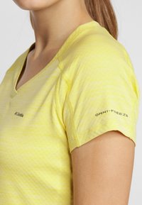 Zbliżenie na osobę w żółtej koszulce sportowej Columbia z dekoltem w serek i napisem "OMNI-FREEZE" na rękawie.