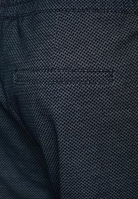 Donkerblauwe stof met een gestructureerd geometrisch patroon, voorzien van een klein zakje aan de achterkant. Het materiaal lijkt lichtgewicht en duurzaam.