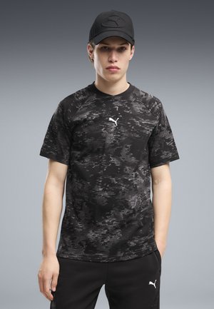 Ung mand iført en sort mønstret Puma t-shirt, sort Puma kasket og sorte bukserstående mod en grå baggrund.