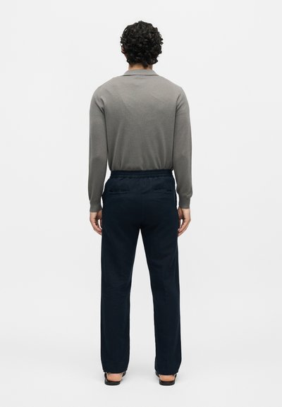 Άνδρας που στέκεται με την πλάτη, φοράει γκρι μακρυμάνικη μπλούζα, σκούρα μπλε παντελόνια με πίσω τσέπες και μαύρα παπούτσια σε λευκό φόντο.