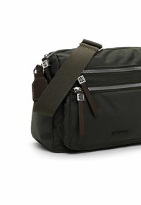 Mørkegrøn nylon taske med sølvfarvede lynlåse, to frontlommer, brune læderdetaljer og en aftagelig justerbar rem.
