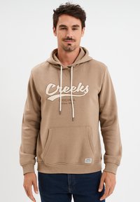 Beige Kapuzenpullover mit Fronttasche, Schnürzug und "Creeks"-Logo in Beige. Verfügt über gerippte Bündchen und Saum, aus weichem Material gefertigt.