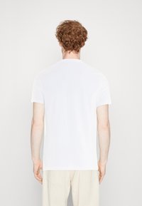 Persona con capelli rossi ricci che indossa una semplice maglietta bianca a maniche corte e pantaloni beige chiaro, vista da dietro su uno sfondo neutro.