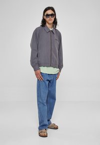 Chaqueta de algodón gris con cremallera frontal y un bolsillo en el pecho, llevada sobre una camiseta verde clara y combinada con unos vaqueros azules holgados y sandalias.
