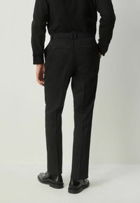 Pantaloni negri, croiți pe măsură, dintr-un material fin, având un design cu picior drept și detalii la buzunarele din spate, asortați cu pantofi negri lustruiți.