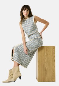 Vrouw zittend op een houten blok, gekleed in een mouwloze tweed top in blauw en beige en een bijpassende rok met beige suède enkellaarsjes.