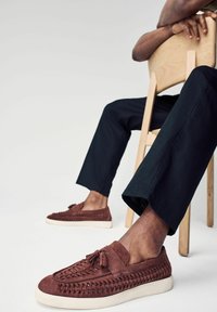 Chaussures en daim marron avec texture tissée décorative et détail de gland, associées à un pantalon sombre, placées sur une chaise en bois contre un fond neutre.