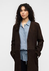 Καφέ μακρύ παλτό από καστόρι με κούμπωμα κουμπιών, σε συνδυασμό με μπλε και λευκό ριγωτό πουκάμισο με γιακά. Λείο ύφασμα και casual σχεδιασμός.