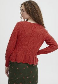 Suéter tejido en rojo con patrones de trenzas y un dobladillo peplum, hecho de hilo texturizado. Lleva puesto sobre una falda de pana verde con estampados florales.
