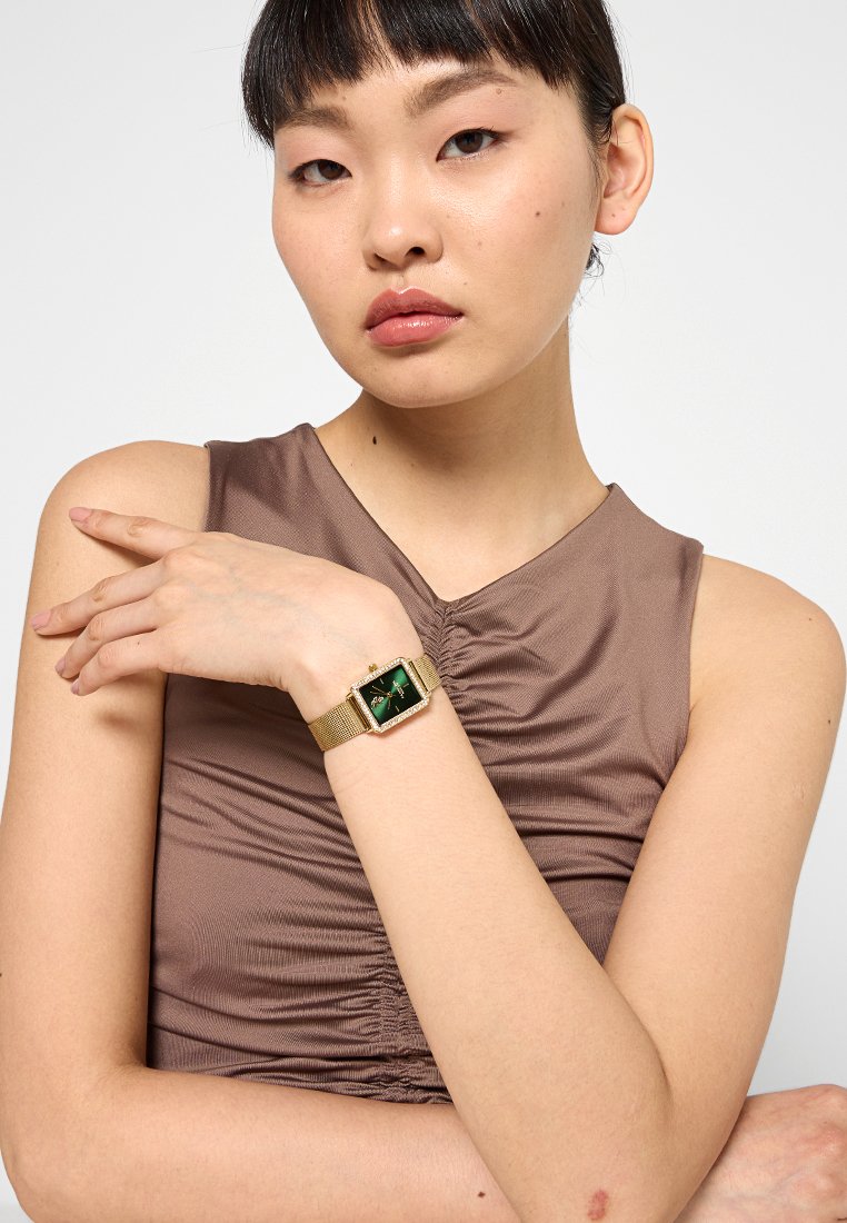Montre-bracelet en mailles dorées avec un cadran rectangulaire vert, ornée d'accents décoratifs et d'un bracelet texturé en or. Modèle posant avec sa main sur le bras.