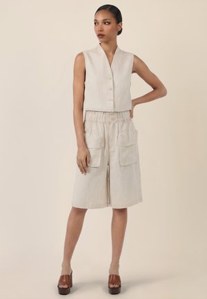 Kvinde i beige, ærmeløs vest med knapper og knælange shorts med store lommer, iført brune platformsandaler, stående med hånden på hoften.