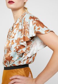 Blomsterbluse med korte flæsekortærmer i lys blå og rustfarver. Har en løs pasform og et sort taljebånd. Glat, satin-agtig tekstur.