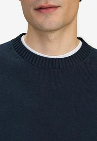 Mörkblå stickad tröja med ribbad halsringning, som har en slät yta och en enkel, klassisk design. Bärs över en vit t-shirt med rund hals.