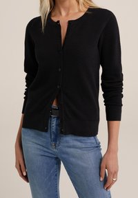 Cardigan nero in maglia con texture, colletto rotondo, chiusura con bottoni frontali, polsini aderenti e orlo leggermente corto, abbinato a jeans blu.