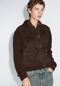 Ung person iført en mørkebrun ruskinds- og strikjakke med forlommer, kombineret med lyseblå denimjeans, som står foran en ensfarvet baggrund.