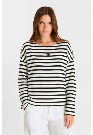 Woman wearing a black and white horizontal striped long-sleeve sweater with white pants, standing against a plain light background.