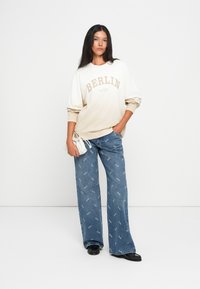 Sweatshirt em creme e bege com o texto "BERLIM", combinado com jeans azuis de perna larga com detalhes de padrão subtil. Modelo segura uma bolsa branca.