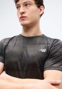 Μαύρο αθλητικό μπλουζάκι με κοντά μανίκια και υφασμάτινο σχέδιο υφής· διαθέτει αντανακλαστικό λογότυπο New Balance στο στήθος. Ομαλό, ελαφρύ ύφασμα.