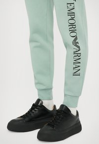 Mentolno zelene taperirane jogger hlače imajo črn logotip "Emporio Armani" ob strani. Ujemajo se z črnimi, masivnimi teniskami in belimi nogavicami.