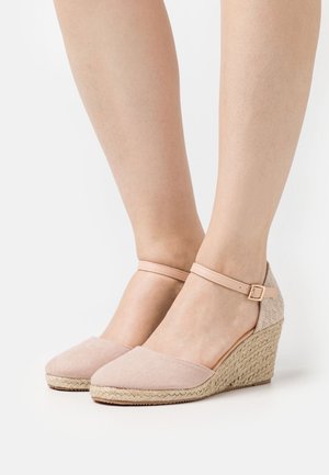 Des pieds chaussés de wedges espadrilles rose poudré avec des brides de cheville et des semelles en jute tressé sur un fond blanc.