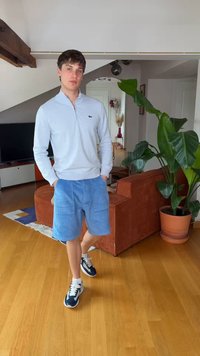 Tânăr stând în interior, purtând un pulover cu fermoar albastru deschis, pantaloni scurți din catifea albastru și adidași, cu o plantă și o canapea în fundal.