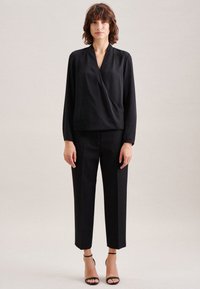 Schwarze Bluse mit drapierter Vorderseite und V-Ausschnitt, kombiniert mit maßgeschneiderten schwarzen Hosen. Das Model trägt schwarze Sandalen mit Riemchen. Glatte Stofftextur.