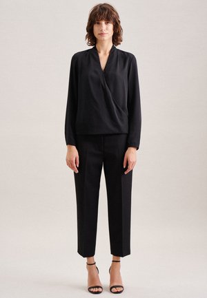 Blusa nera con scollatura a V e drappeggio frontale, abbinata a pantaloni neri su misura. La modella indossa tacchi a spillo neri. Tessuto dalle linee morbide.