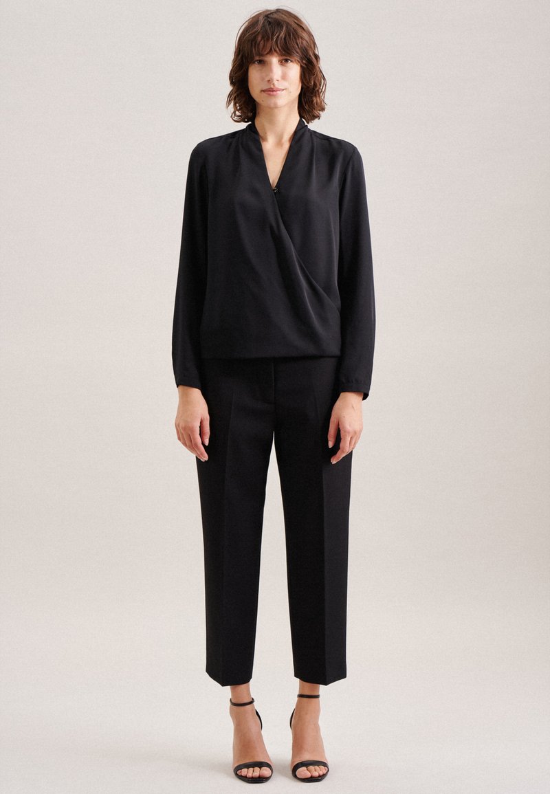 Schwarze Bluse mit drapierter Vorderseite und V-Ausschnitt, kombiniert mit maßgeschneiderten schwarzen Hosen. Das Model trägt schwarze Sandalen mit Riemchen. Glatte Stofftextur.